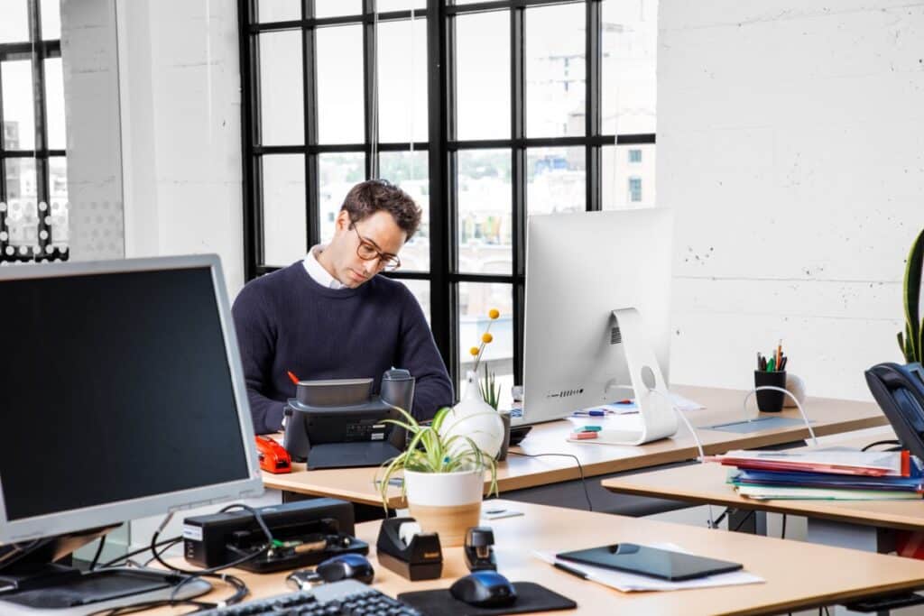 Male office worker standing at desk with desktop computer. Blank screen shown on monitor in foreground.