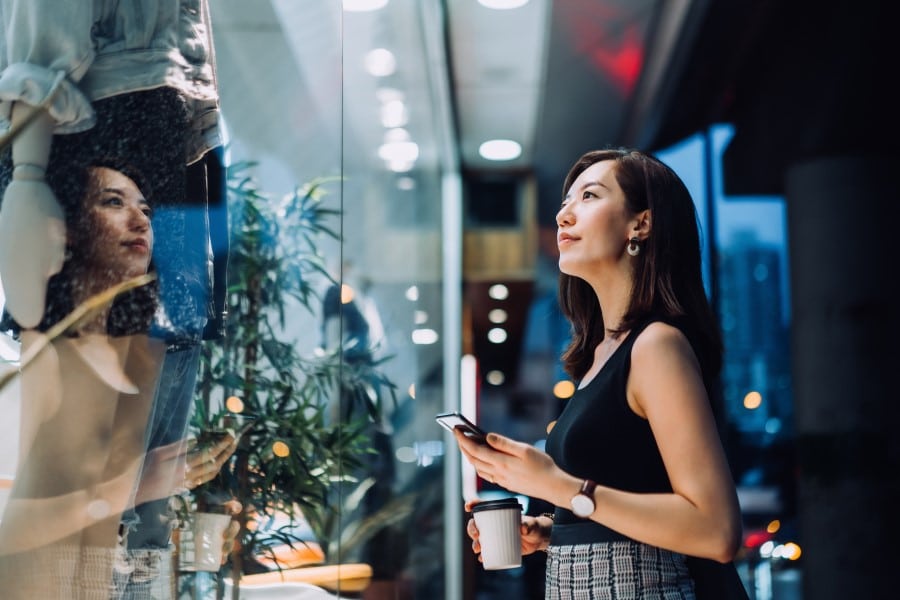 Cheerful young Asian person holding a cup of coffee, checking their smartphone while standing outside a boutique looking at shop window in the evening in the city