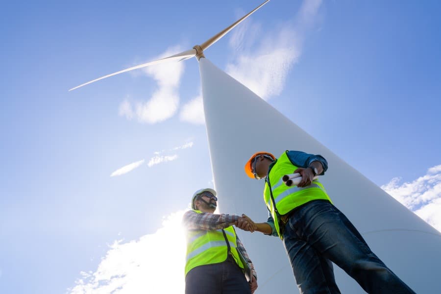 Two field workers shake hands beneath a tall wind turbine