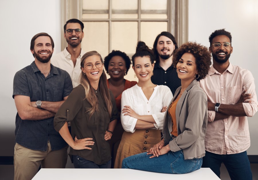 8 professionals of varied ethnic and age backgrounds in casual attire grouped together, smiling at the end of a conference room.