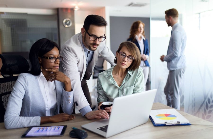 Two female finance professionals sitting at a desk looking at laptop and tablets with financial graphs on them and one male finance professional leaning over behind them navigating something on the laptop.