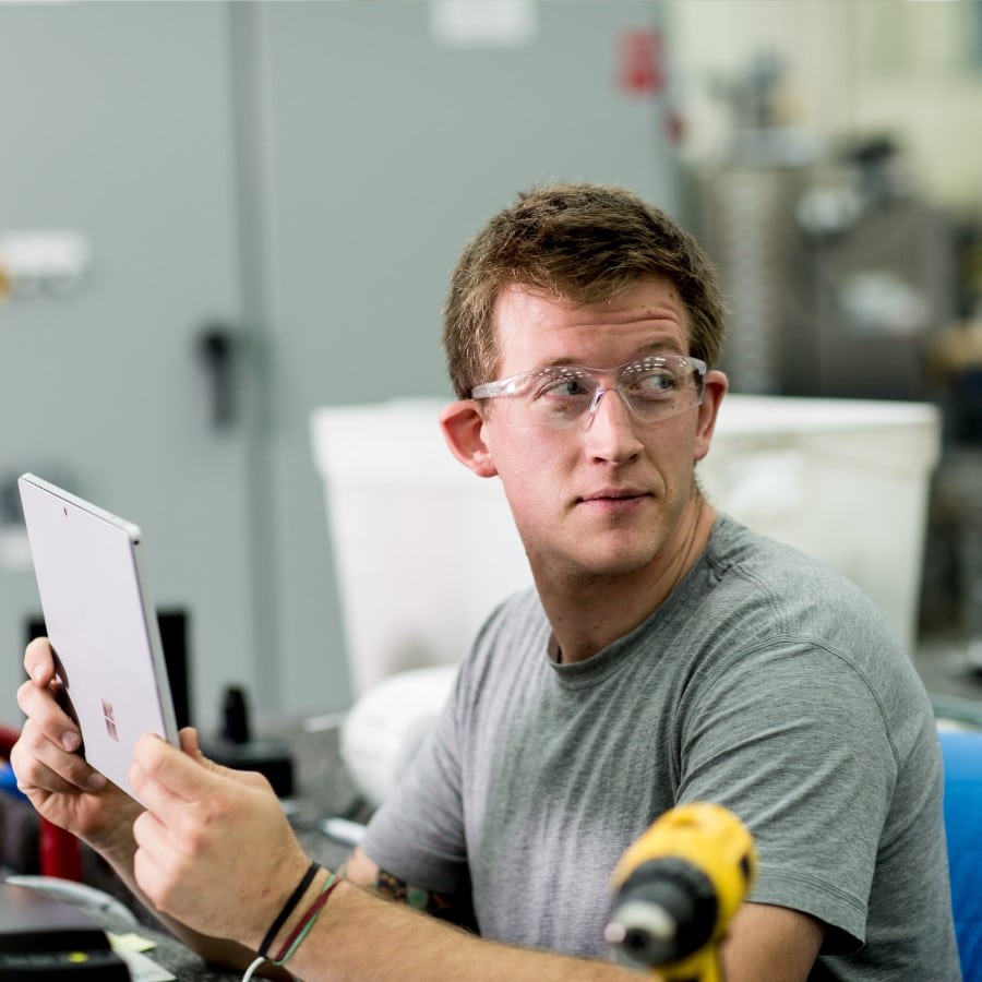 Male factory worker sitting at desk in manufacturing plant. He is wearing safety glasses and looking over his left shoulder while holding a Surface Pro (screen not shown).