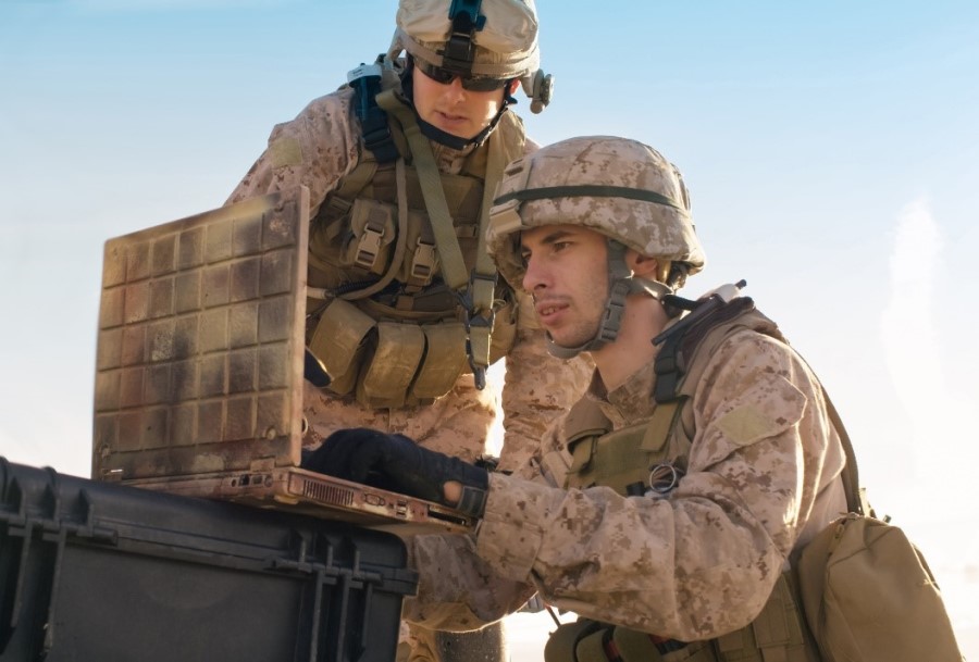 Two men in tan camo military uniforms in the field looking at a computer.