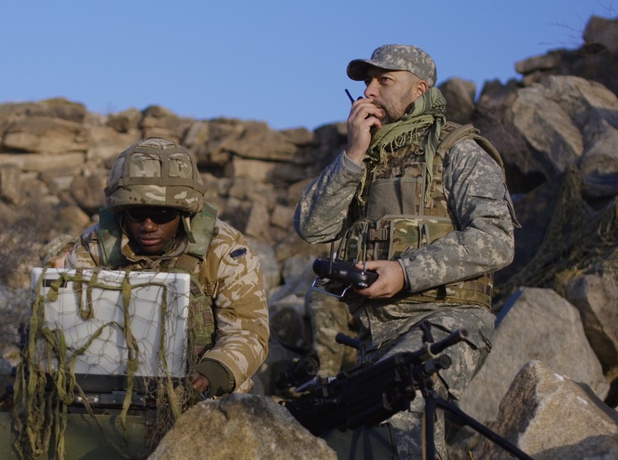 One man in a military uniform with a helmet sitting looking at a laptop out in the field and another man in military uniform talking on walkie talkie.