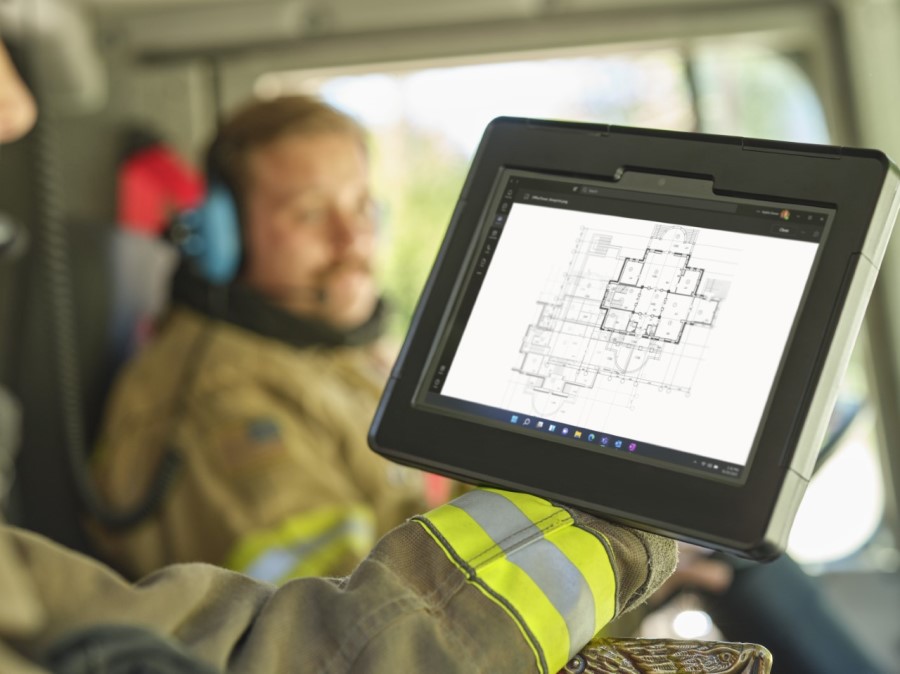 In the forefront, the arm of a firefighter sitting in the passenger seat of a firetruck holding a Surface tablet to look at blueprints of a building and a male firefighter in the background in the driver's seat with a headset on.