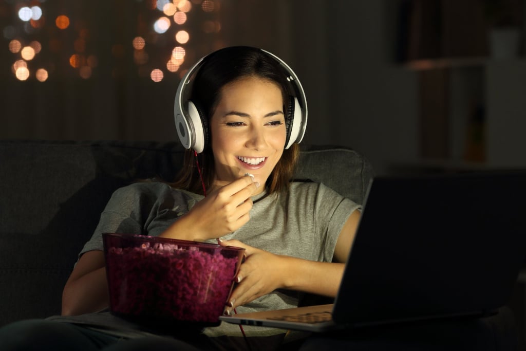 A woman smiling and wearing headphones watching a screen and eating popcorn