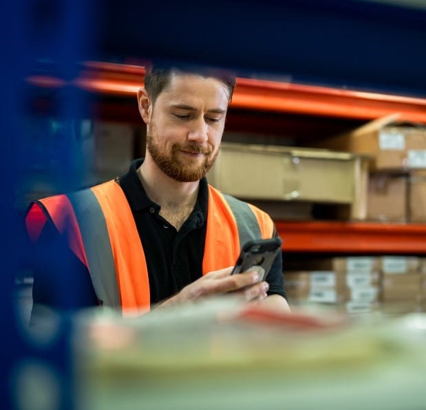 Man wearing an orange vest in a manufacturing or warehouse who appears to be doing inventory or looking at something on the shelf and messaging about it on his phone in Microsoft Teams.