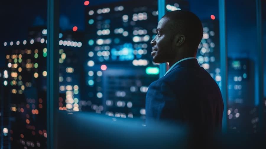 A businessman in the financial services industry looks out an office window, the lights of nearby buildings in the background, as he reflects on the new year ahead.
