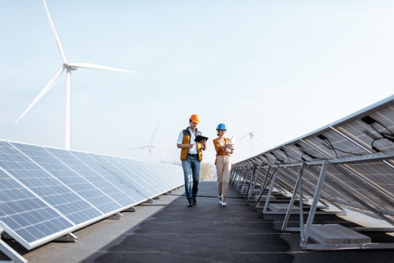 Field workers with tablets walking near solar panels and wind turbines