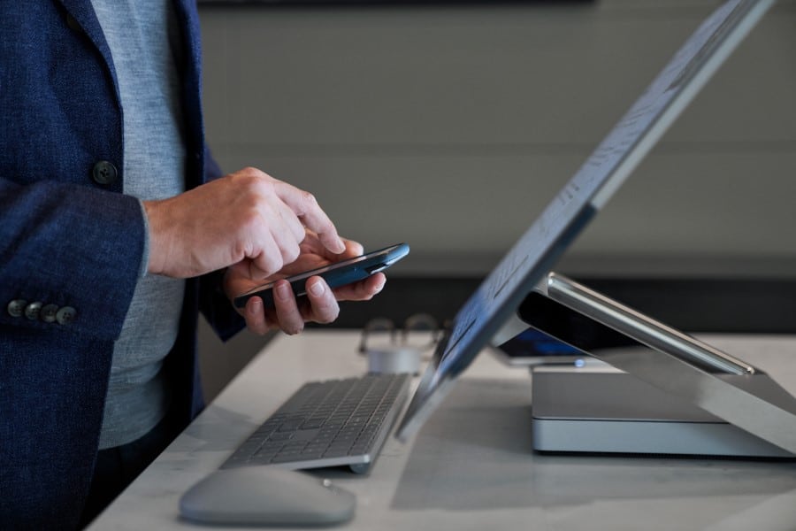 Side view close-up of a man typing on his phone while standing behind a Microsoft Surface Studio.