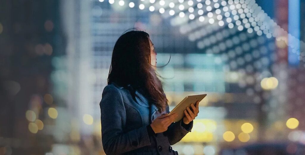 Person holding tablet looking back toward a lit up city.