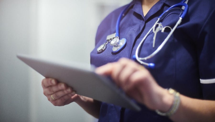 Female surgeon typing on digital tablet in hospital or surgery. She is wearing a dark blue nurse’s top and has her stethoscope around her neck. She is looking at her patients records on her digital tablet