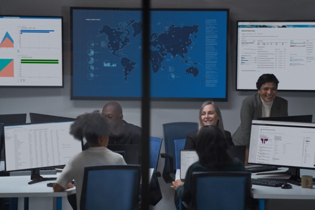 A group of people sitting at a table in a meeting room with many monitors