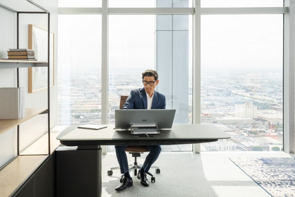 A man sitting at an office desk working on a Surface Studio 2 Plus and a Surface Pro 9 on the desk next to him. No screen showing.