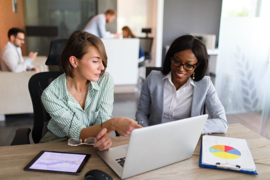 Two businesswomen are in the office using laptop, digital tablet and chart papers while working together.